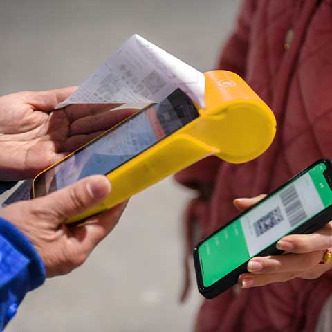 This closeup image shows a driver making a smartphone payment at a gas station. The driver holds her phone up to a device held by a gas station worker.  