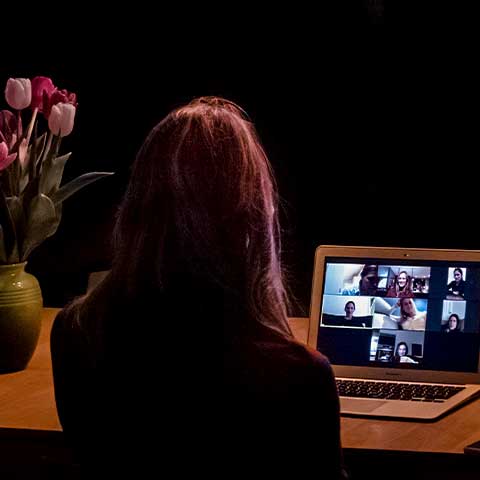 A woman on a Zoom call. She sits in a dark room with her back to the camera. Her laptop is open on the table in front of her and other Zoom callers are shown on her computer screen. There is a vase of flowers on the table. 
