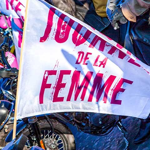 A picture of women celebrating International Women's Day in Paris, France with a flag mounted to the back of a motorcycle showing the words "Journee de la Femme".