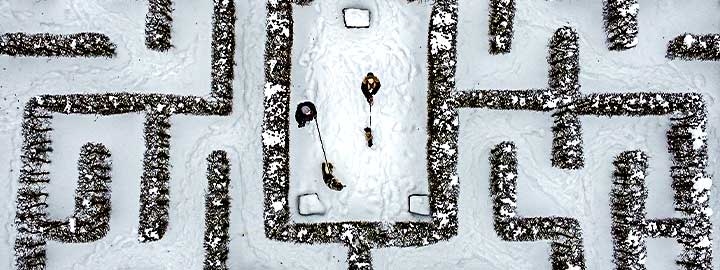 This is an aerial view of a snow covered garden maze in Gelsenkirchen, Germany. Two people walking dogs are at the center of the maze. The center is surrounded by  snow-covered pathways bordered by shrubbery.