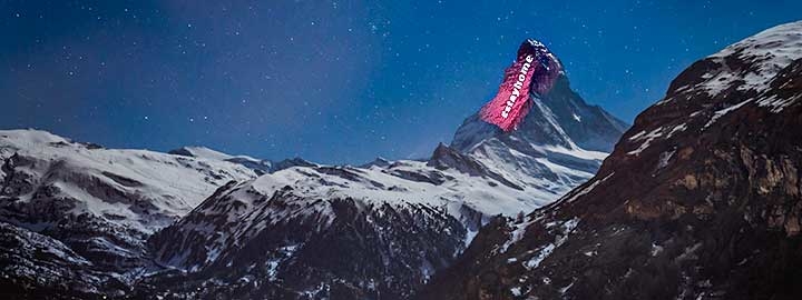 A nighttime image shows the lights of Zermatt, Switzerland in the foreground. Beyond Zermatt, the alps rise. The Matterhorn is illuminated by a light installation meant to foster hope during COVID-19. The message projected on the mountain says "#StayHome".