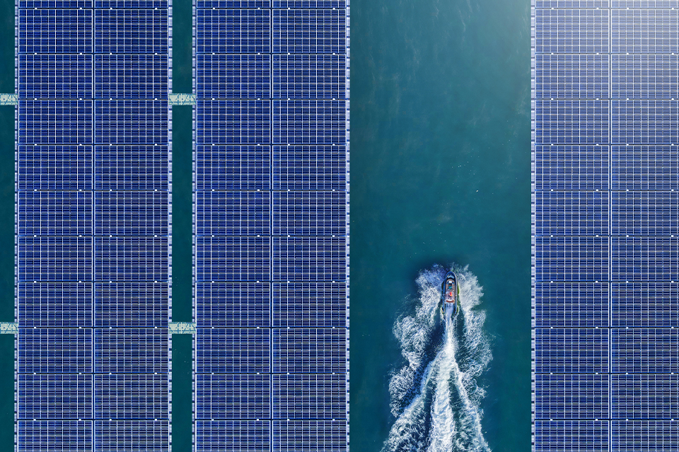 Bird’s-eye view of three solar panels floating in a dam, providing a source of renewable energy. There are a couple of people riding a jet ski between the two solar panels on the left side and the one on the right. 