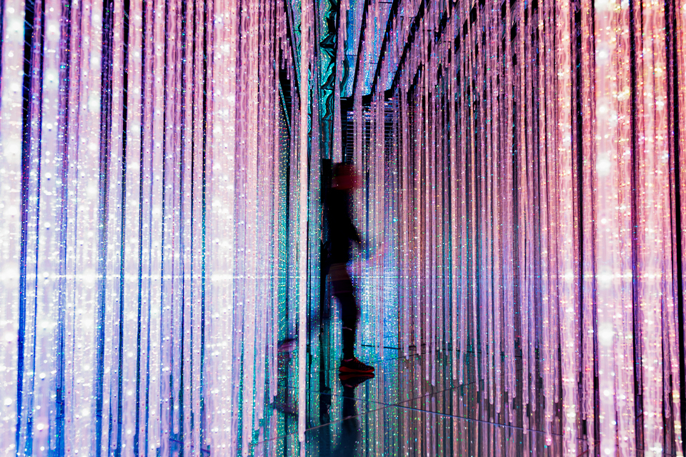 A blurry male figure walks through colorful strips of bright, sparkling lights. The lights cascade from the ceiling to the floor.