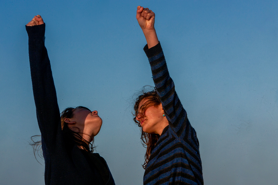 Two teenage girls facing each other raise their fists against a late-afternoon blue sky. One girl, left, looks up, while the other, right, looks into the distance. The wind blows their hair. 