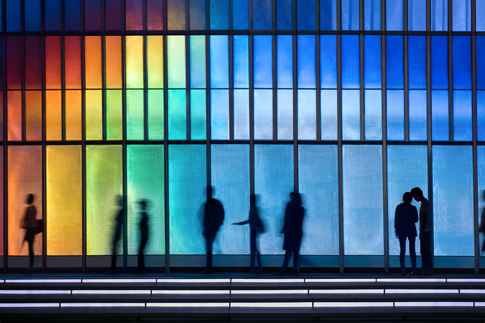 Silhouettes of individuals walking past an LED-lit façade of a building in Hong Kong, China, showcasing vibrant colors that constantly change. There are also two people talking in front of the building.