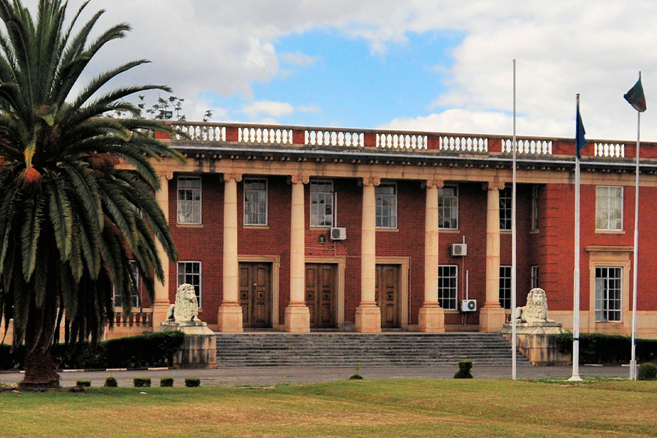 A distant view of the Supreme Court of Zambia, in the country's capital of Lusaka. The red-brick, two-story building features white columns along the main facade and a pair of white lion sculptures enclosing the wide entrance steps. A large palm tree stands to the left, and three flagpoles to the right.