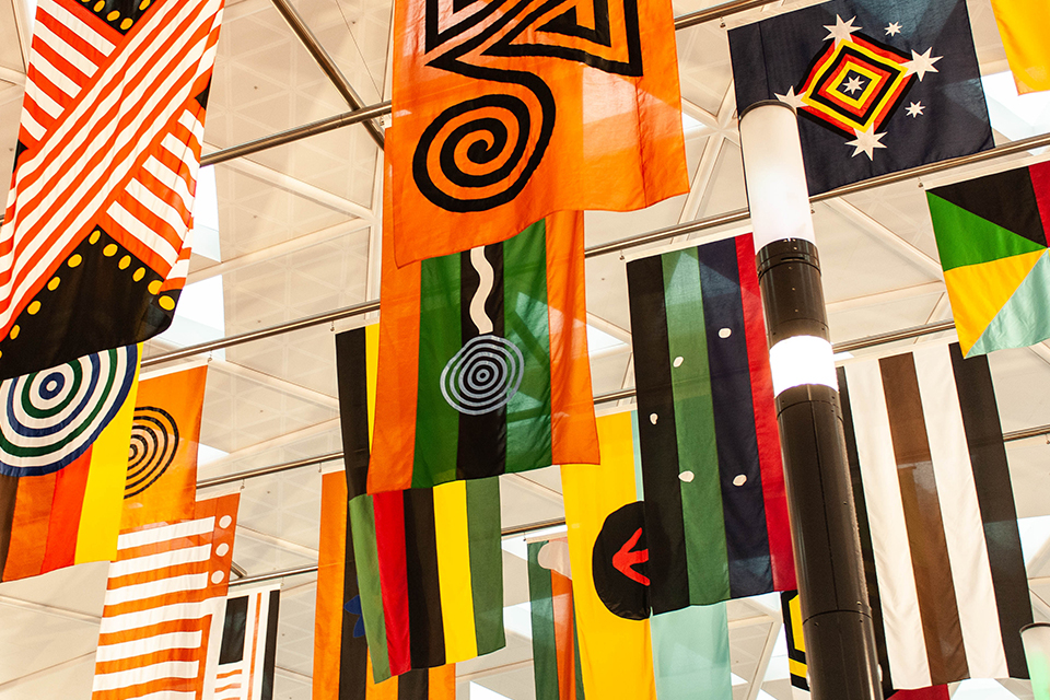 A close-up view of 28 colorfully decorated flags that form the art installation "United Neytions," which hangs in Sydney Airport. Each flag represents one of the original Aboriginal nations and is decorated in varying combinations of stripes, dots, spirals, diamonds and stars, in a color palette of orange, green, black, red, yellow and white. The work is by artist Archie Moore, a member of Australia's Kamilaroi Indigenous nation. 