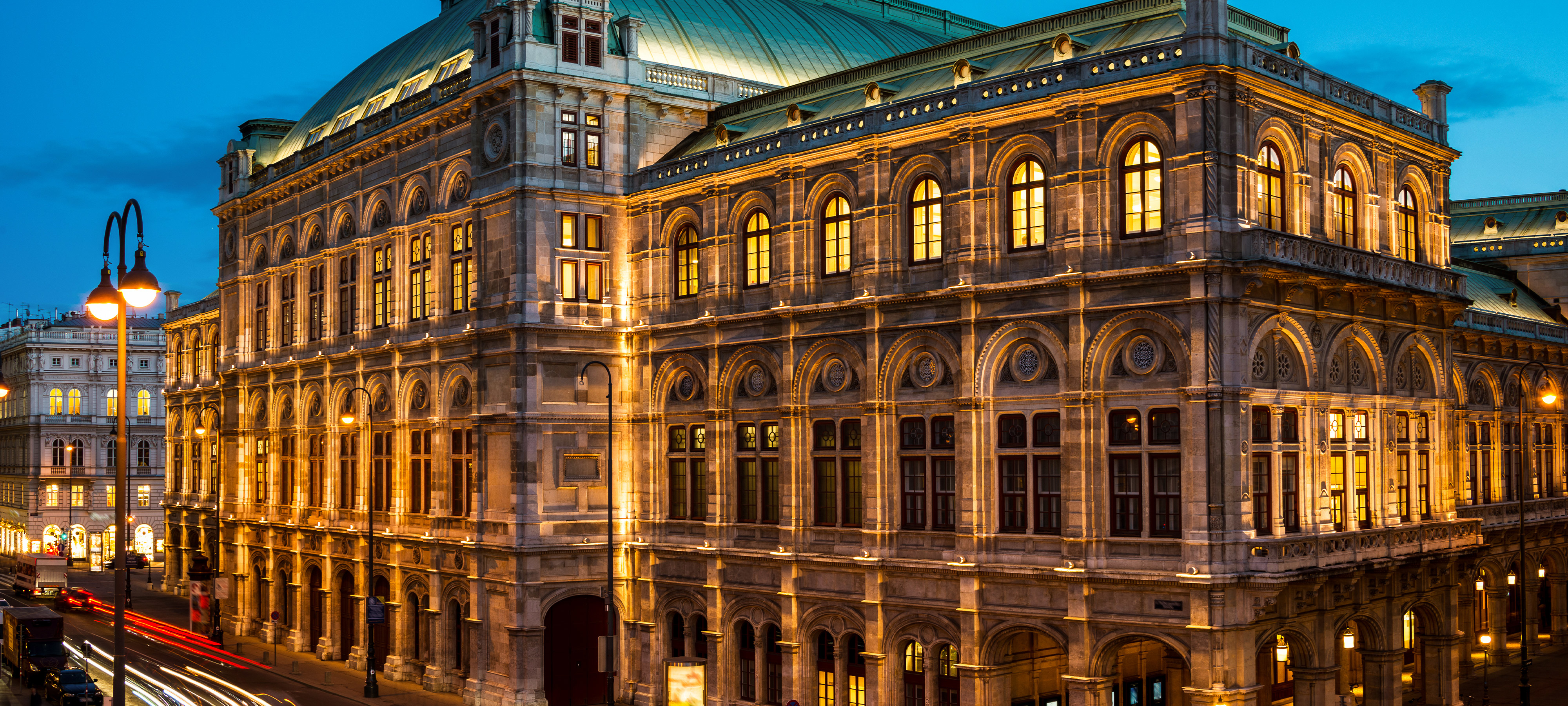 A nighttime view of the Wiener Staatsoper (Vienna State Opera) in Vienna, Austria. The Neo-Renaissance building is warmly lit, inside and out. Light from traffic appears as red and white streaks in the left foreground.