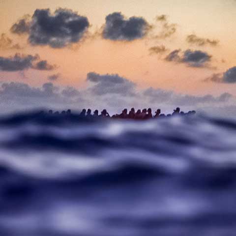 This image, taken from a distance, shows migrants from Libya in a boat on the water. They are waiting for help from a Spanish organization. Water takes up most of the image. Far off, the outlines of the migrants sitting in their boat appear against a sky dotted with clouds. 