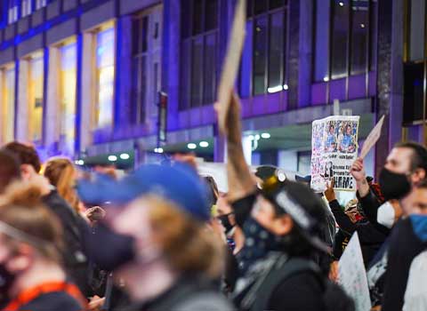A nighttime image shows a crowd of masked protesters in New York City. They are holding signs and demonstrating against the killing of Breonna Taylor by police. There is an office building in the background. 