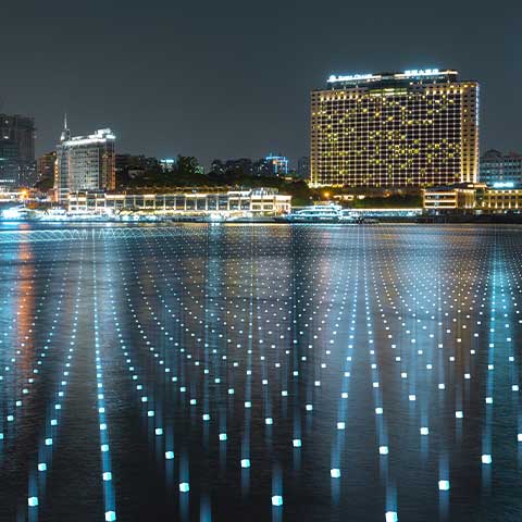 This photo shows the distant skyline of a Chinese city against a dark sky. Lights are in on in some of the buildings’ windows. Rows of lights spaced at even intervals take up the entire foreground of the image. These are smart city grids. 