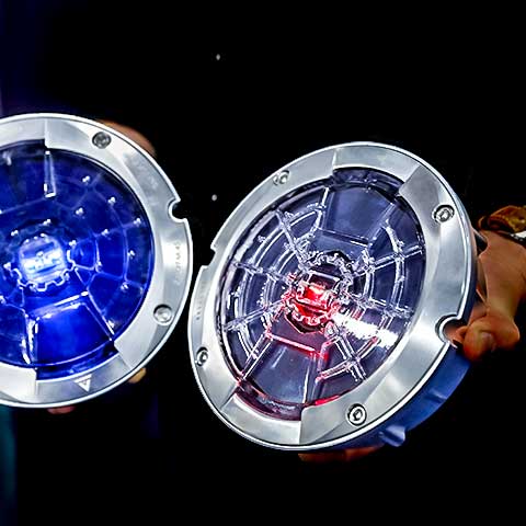 A closeup image of two smart road studs, which can monitor traffic and road conditions. Each stud is circular with a metallic rim and light at its center. They appear against a dark background. An outstretched hand holds one of the studs. 