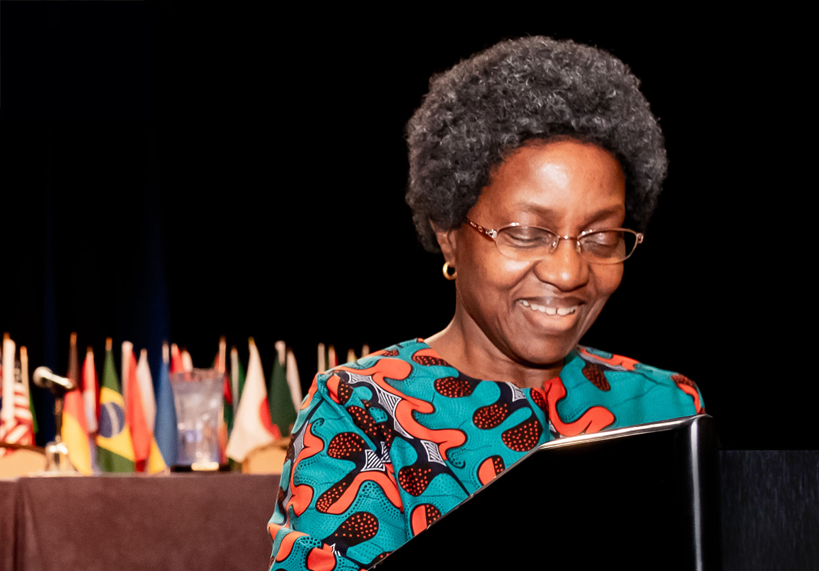 Close-up view of Judge Florence Mumba standing at a podium to accept the White & Case Distinguished Jessup Alumni Award at the White & Case Jessup International Rounds in April 2023, in Washington, DC. She is wearing a colorful blouse and looking down and smiling. A row of flags decorates the stage behind her.