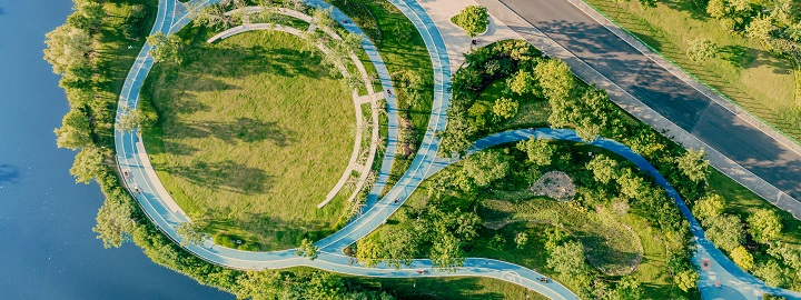 An aerial view of a city park at sunset in China. Paths wind gently through a verdant landscape, joining at the top left to form a circle enclosing an open, grassy space. Water is visible to the left, and a main road cuts through the upper right.