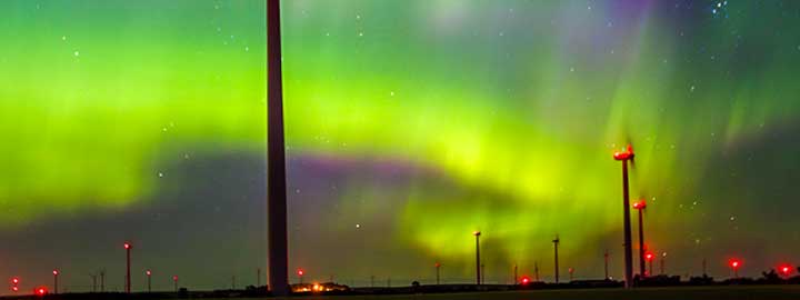 Blurred photo showing the vivid colors of the Northern Lights in the sky over a wind farm in North Dakota. Tall wind turbine towers are outlined against the sky. Their blades are not clearly visible, but there is a bright light at the top of every tower.  