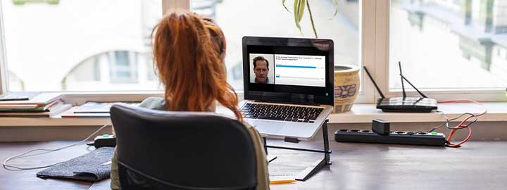 An image of a woman viewing a webinar at her desk. The back of her head faces the camera and we see the webinar on her laptop screen in front of her. Beyond her laptop is a window, with a plant sitting on the sill. 