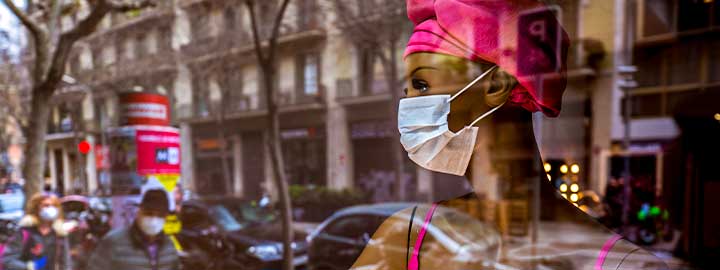 A photograph of the reflection of a female mannequin in a shop window wearing a pink dress and a white face mask as people walk past on the sidewalk in Barcelona, Spain, March 2020.