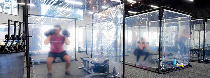 A photo of two people exercising with weights behind clear plastic sheets in individual workout pods to observe social distancing in Redondo Beach, California in June 2020.