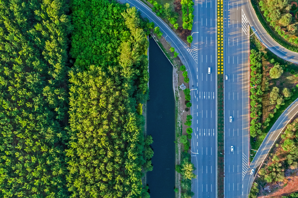 An aerial view of a mostly empty eight-lane highway in Beijing. A median divides the highway. One vehicle travels in one direction, and two travel in the other. Groves of trees surround the highway.
