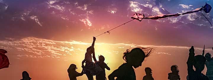 Syrian children are silhouetted against the sky at a refugee camp near the Turkish border. One child is flying a kite. Beyond the children, the sun is low in the sky and there are clouds above it. 
