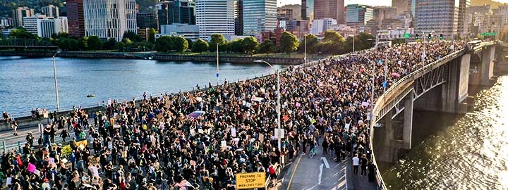 An aerial view of a densely packed crowd of people crossing Morrison Bridge in Portland, OR. They are protesting the death of George Floyd while in police custody. Sunlight reflects off the water on either side of the bridge, which is meant for vehicular traffic. In the distance, beyond the bridge, are trees and tall city buildings. 