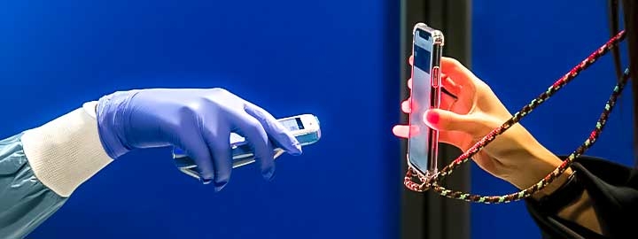 A closeup image of two hands holding smartphones. The gloved hand of a medic extends to scan a phone being held by a traveler at Athens International Airport prior to COVID-19 testing. The image includes the masked lower portion of the traveler’s face.