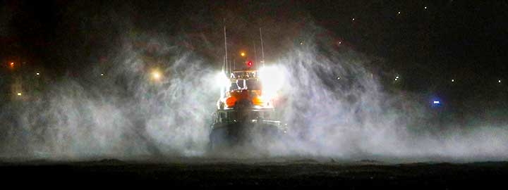 A nighttime image of a fishing boat moored at a port in the Azores during Hurricane Lorenzo. The lights of the boat and some of its outline appear at the center of the image. Heavy rain and winds create the appearance of fog surrounding the boat. The rest of the image is dark except for stars in the sky. 