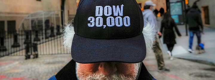 A closeup image of a baseball cap that says ”DOW 30,000“ worn by a trader standing outside the New York Stock Exchange. The man’s eyes are hidden by the bill of the cap. He has gray hair beneath the cap and a gray beard and mustache. The scene behind him is blurred and includes passersby.