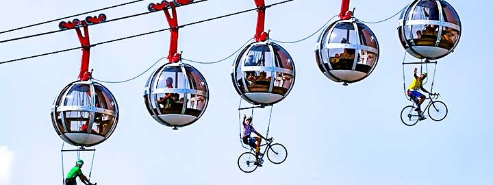 This image shows five globe-shaped gondola cars ascending left to right, suspended from a cable, in Grenoble, France. Performers dressed as cyclists and seated on bicycles are suspended beneath three of the gondola cars.