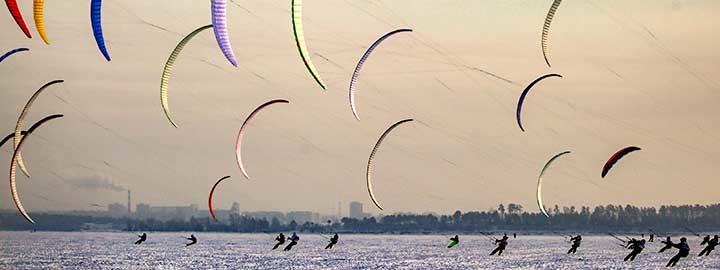 An image of colorful C-shaped kites in the air above snow-covered ground in Novosibirsk, Russia. The kites are held by people on skis in the snow.