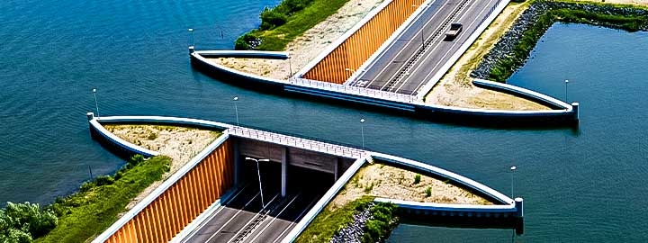 A daytime overhead image of an aqueduct in Veluwe Lake in Flevopolder, Holland. A multi-lane road, surrounded by water, bisects the image. 