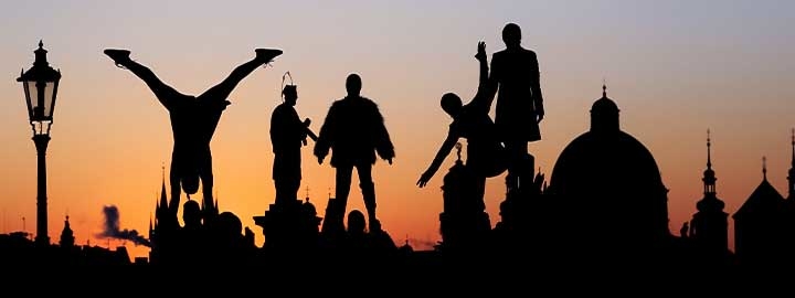 Street artists perform, and statues of people and the dome of a building are silhouetted against the sky in Prague, Czech Republic.