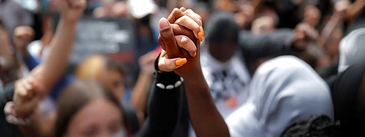 A closeup image of a Black person and a white person holding hands at a protest against the death of George Floyd while in police custody. The hands are raised in the air above a blurred crowd in Nantes, France.