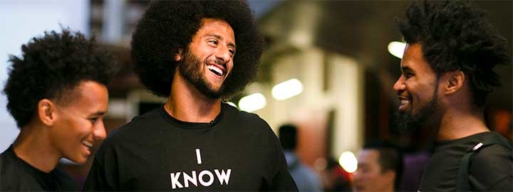 Activist and former NFL quarterback Colin Kaepernick is talking with two teens at a community center in Oakland, CA. They are at an event held to educate youth about their rights, especially when dealing with the police. Kaepernick stands in the middle of the group. He and the teens are smiling and wearing t-shirts that say ”I KNOW MY RIGHTS.” Behind them, and blurred, are lights and other people. 