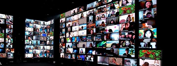 This image shows two artists in a dark room surrounded by walls of video screens that show the members of their audience. They are participating in a remote music festival from a studio in Bangkok.