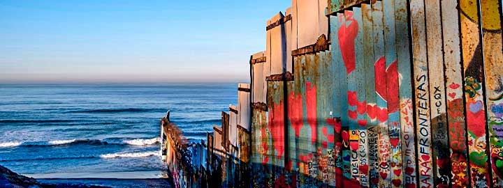A photo of the US-Mexico border fencing at Playas de Tijuana, Mexico, running down a hill to the Pacific Ocean. The fence is made of rusted, patchy metal and covered with graffiti.
