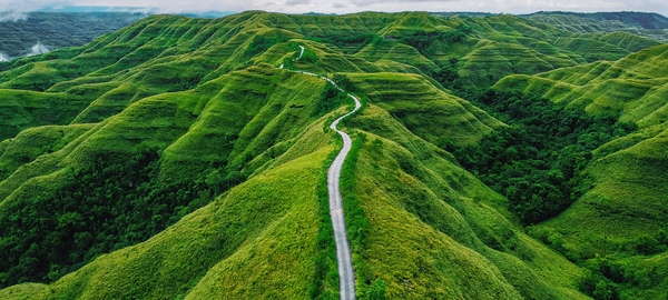 Hills, covered with vegetation, stretch toward the horizon, where clouds and sky are visible. At the center of the image, a winding road cuts through the hills heading in the direction of the horizon. 