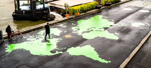 A person takes a daytime walk across a bridge that crosses the Seine River in Paris. The surface of the bridge features a large, vividly colored world map against a dark background. Another person on the bridge stands beside a bicycle.