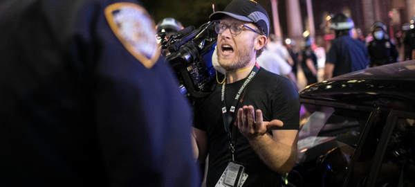 A journalist holding a TV camera on his right shoulder faces a police officer seen from behind while a protest is going on in the back.  The journalist is reminding the officer that the press are considered "essential workers" in regard to curfews related to protests.