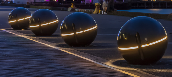 Four large metal spheres, each bisected by a strip of light, sit on the boardwalk along the harbor in Wellington, New Zealand. Water and the skyline are behind them. 