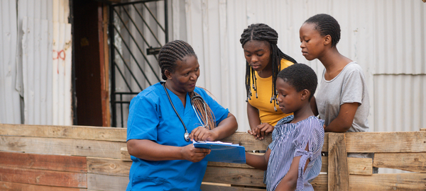 A community nurse, left, shares information on a clipboard with three teenage girls, right. The group stands by a wooden fence in front of a shack in an informal settlement in Africa.