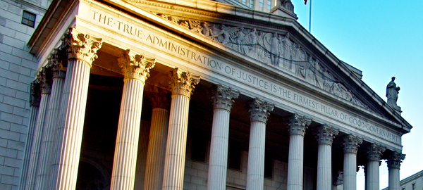 A view of the portico of the New York County Supreme Court, in lower Manhattan in New York City. Above its columns, the portico is engraved with the motto, "The true administration of justice is the firmest pillar of good government." Sun shines on the top left corner.