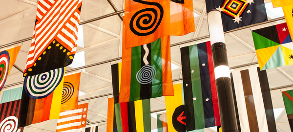 A close-up view of 28 colorfully decorated flags that form the art installation "United Neytions," which hangs in Sydney Airport. Each flag represents one of the original Aboriginal nations and is decorated in varying combinations of stripes, dots, spirals, diamonds and stars, in a color palette of orange, green, black, red, yellow and white. The work is by artist Archie Moore, a member of Australia's Kamilaroi Indigenous nation. 