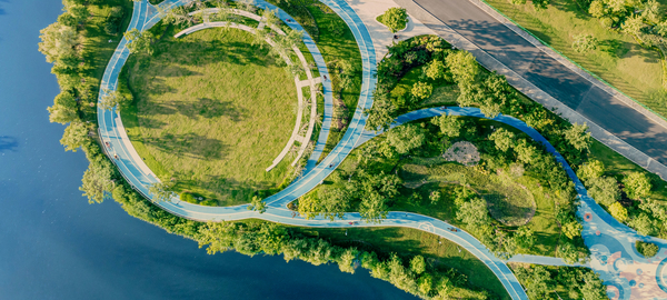 An aerial view of a city park at sunset in China. Paths wind gently through a verdant landscape, joining at the top left to form a circle enclosing an open, grassy space. Water is visible to the left, and a main road cuts through the upper right.