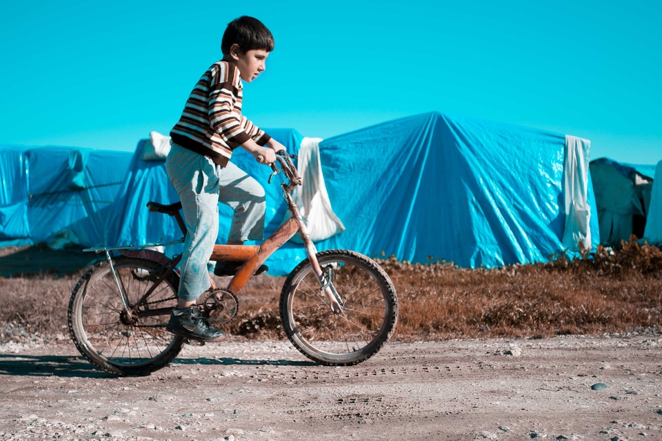 A child in a striped shirt and light-colored pants rides a bicycle along a dirt road during the day, passing in front of a refugee camp. The camp consists of several blue tents.