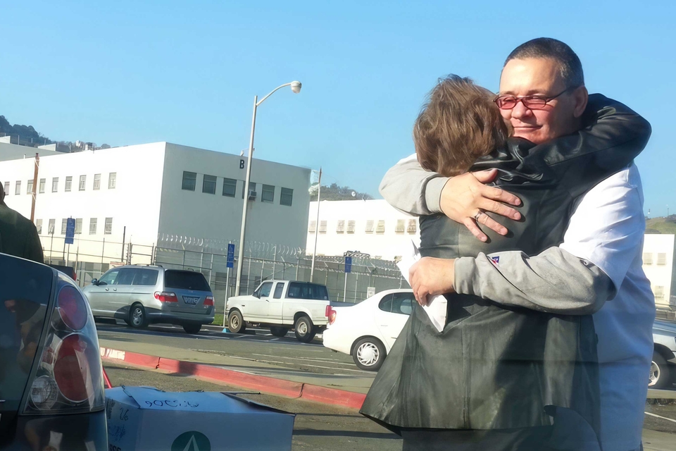 Two people, one n wearing glasses and a short-sleeve shirt over a long-sleeve shirt, embrace in a parking lot outside a prison during the day. The lot, separated from the white prison by a fence, contains several cars and SUVs.