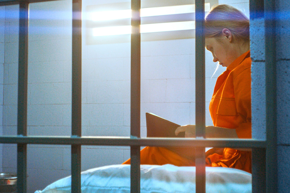In a small prison cell, a woman in an orange jumpsuit sits against a brick wall reading a book as sunlight shines through the window vents.