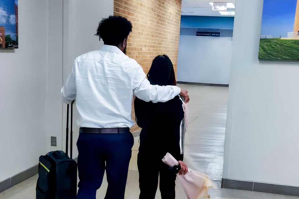 After being reunited, a father places his right arm over his daughter's shoulders as they exit the waiting area in an airport. His left hand grips the handle of a carry-on suitcase.