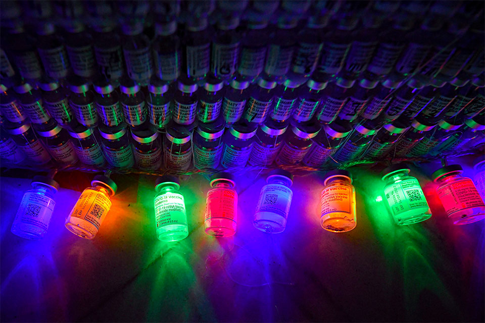 Bright, colorful electric lights inside empty COVID-19 vaccine containers shine beside more empty containers, which are stacked to resemble a Christmas tree 