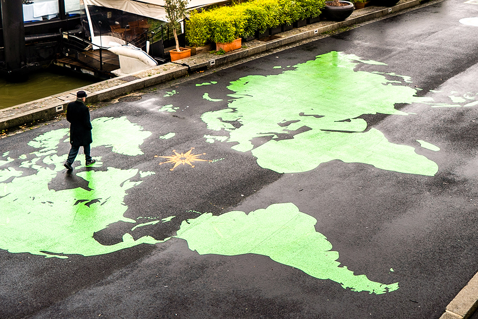 A person takes a daytime walk across a bridge that crosses the Seine River in Paris. The surface of the bridge features a large, vividly colored world map against a dark background. Another person on the bridge stands beside a bicycle.